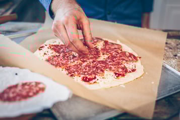 Die Pizza liegt auf einem Backpapier bereit und wird mit Salami belegt.
