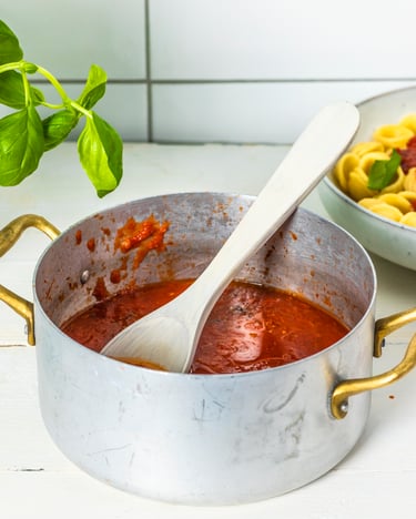 Topf mit Tomatensauce, dahinter steht Pasta und ein Topf mit Basilikum
