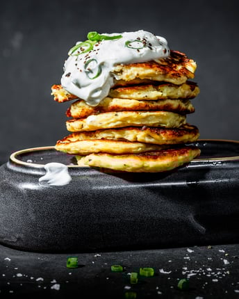 Ein Stapel von goldbraun gebackenen Kartoffel-Pancakes mit einem Klecks Crème fraîche on top und Frühlingszwiebeln vor dunklem Hintergrund