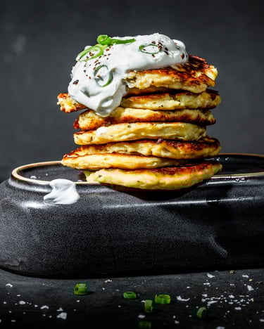 Ein Stapel von goldbraun gebackenen Kartoffel-Pancakes mit einem Klecks Crème fraîche on top und Frühlingszwiebeln vor dunklem Hintergrund