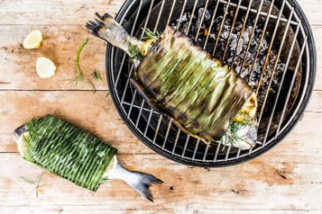 Ein Fischpäckchen liegt auf einem Rost auf einem Grill, das zweite Päckchen daneben.