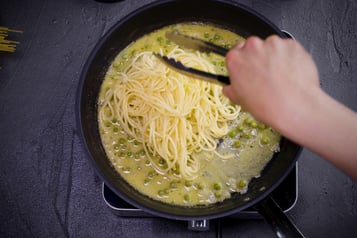Spaghetti werden mit einer Zange mit der Erbsen-Weißweinsoße in der Pfanne vermischt.