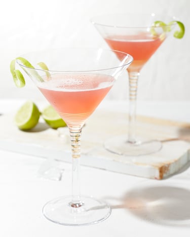 Zwei Martinigläser mit rötlichem Cosmopolitan und Limettenschale in einem hellen Küchensetting auf einem weißen Untergrund mit Schneidebrett, halbierter Limette und Cocktailshaker im Hintergrund