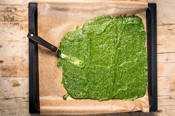 Spinatmasse ist auf einem Backblech mit Backpapier verstrichen. Darauf liegt ein Spachtel