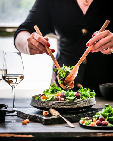 Zwei Hände richten mit Holz-Salatbesteck einen Feldsalat mit Weintrauben an. Daneben steht ein Teller mit Salat und zwei Gläser Weißwein.