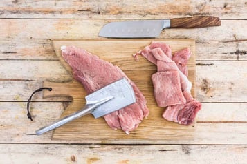 Einplattiertes Schnitzel auf einem Holzbrett. Darauf liegt ein Plattiereisen. Rechts daneben unplattierte Schnitzel. Darüber liegt ein Küchenmesser mit Holzgriff