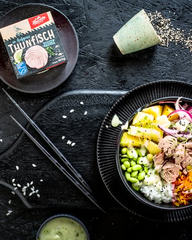 Farbenprächtig sticht die bunte Bowl vor dem schwarzen Hintergrund hervor. Edamame, Ananas, Reis, Zwiebeln, Möhre, Blattspinat und Thunfisch sind ringsum angerichtet.