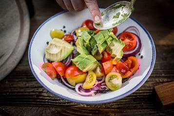 In eine Schüssel mit Tomatenvierteln und roten Zwiebelringen werden Avocadowürfel von einem Esslöffel mit einem Finger geschoben