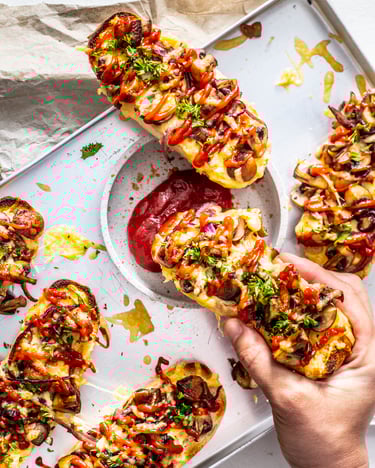 Mehrere dick belegte gebackene Baguettes mit Pilzen, Käse und Ketchup liegen auf einem silbernen Blech. Eines wird gerade in Ketchup gedippt.