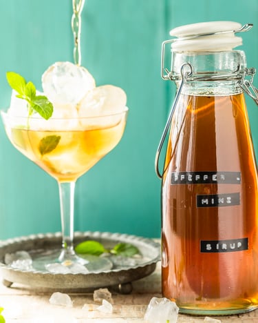 Eine Flasche mit selbstgemachtem Pfefferminzsirup steht neben einem Glas, das gefüllt ist mit Eiswürfeln und verdünntem Minzsirup.