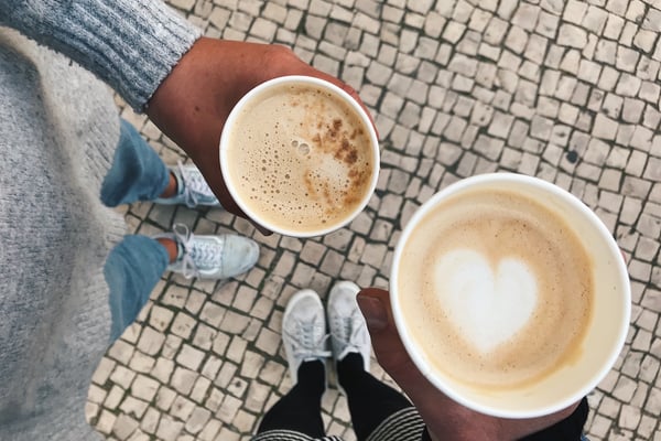 Zwei Becher Kaffee aus der Vogelperspektive fotografiert, darunter die Beine der Personen, die die Kaffees halten
