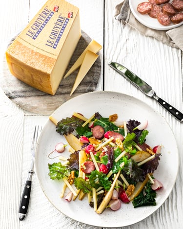Ein weißer Teller, der mit einem Salat aus Grünkohl, Gruyère-Käsestreifen, Radieschen und Mettwurst gefüllt ist, stehen neben einer Schale mit dünn geschnittener Mettwurst und einem Stück Gruyère auf einem weiß gebeizten Holzuntergrund