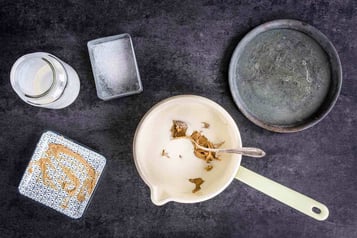 Erdnussbutter wird in einen weißen Topf mit Sahne eingerührt, daneben eine rechteckige Dose mit Zucker und ein runder Metallteller
