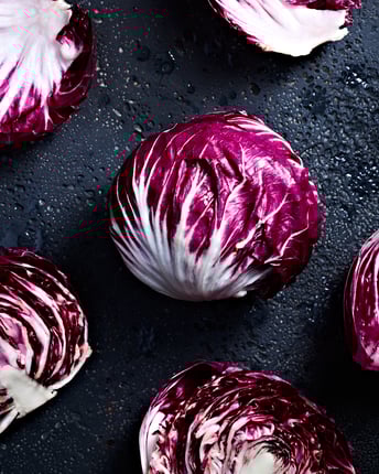 Ein ganzer, ein halbierter und mehrere lose Blätter von Radicchio-Salat nebeneinander auf dunklem Untergrund