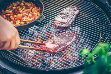 Auf einem großen Grill werden zwei Scheiben Ribeye-Steak gegrillt. Eine Hand mit Holzzange wendet eins der Steaks. Danebenm eine Pfanne mit Tomaten-Kartoffel-Würfeln.