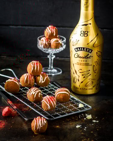 Braune Pralinen mit weißer und roter Creme als Dekoriert sind serviert in einem Glaskelch und auf einem Metallgitter auf einem schwarzen Tisch. Daneben steht die edle Flasche Baileys Chocolate Deluxe in festivem Gold