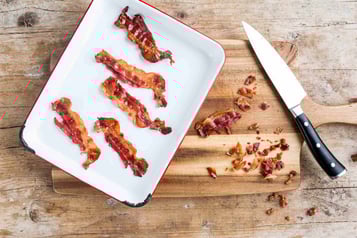 5 Stücke Bacon liegen auf einem Teller auf einem Holztisch, daneben ein Messer und kleine Stücke Bacon