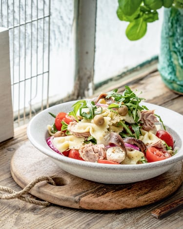 Nudelsalat mit Mozzarella, Thunfisch und anderen Zutaten in weißem Teller vor einem hellen Fenster