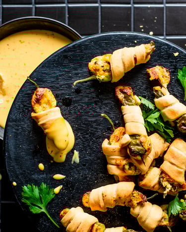 Ein dunkler Teller mit mehreren kleinen von Pizzateig umwickelten Pimientos Poppers und frischer Petersilie in Topshot von oben neben einer Schale mit gelbem Käse-Dip auf schwarzem Fliesenuntergrund