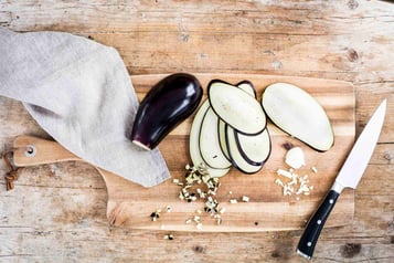 Auf einem Holzbrett liegt eine Aubergine, daneben geschnittene Auberginenscheiben