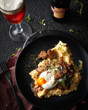 Ein schwarzer Teller mit cremiger Parmesan-Polenta, in Bier sautiertenPilzen und einem pochierten Ei neben einem Glas mit Duckstein