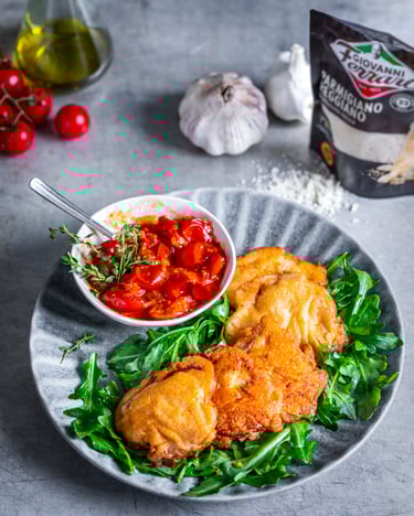 Piccata alla milanese mit Tomatenchutney auf Rucola, daneben eine Tüte geriebener Parmesan