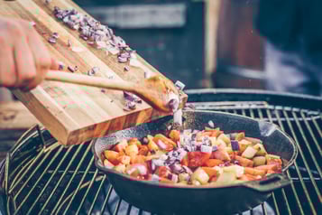In eine Pfanne auf einem Grill mit Tomaten- und Kartoffelwürfeln wird rote, gewürfelte Zwiebel gegeben.