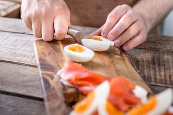 Auf einem Holzbrett wird ein gekochtes Ei mit einem Messer geschnitten.Unscharf im unteren Bildrand ein Brot mit Räucherlachs und Ei