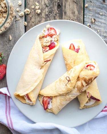 Drei zusammengerollte Pfannkuchen mit einer Füllung aus Erdbeeren, Cheesecake und Kekskrümeln liegen auf einem hellen Teller neben frischen Erdbeeren auf einem Holzuntergrund.