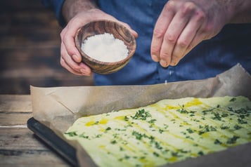 Focaccia liegt auf einem Backblech und wird mit Salz betreut