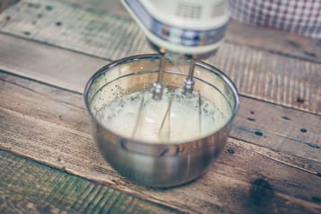 Zuckersirup wird in einer Metallschüssel mit einem Handrührgerät aufgeschlagen. Die Schüssel steht auf einem Holzuntergrund