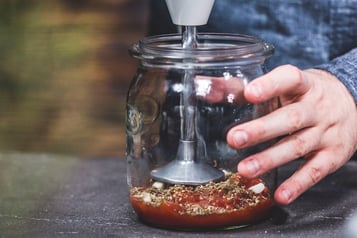 In einem Glasgefäß wird Tomatensauce mit Kräutern mit einem Pürierstab püriert.