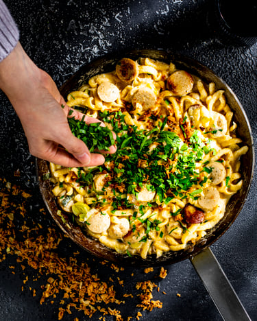 Spätzle mit Weißwurst in einer schwarzen Pfanne, darauf wird von einer Hand Petersilie gestreut