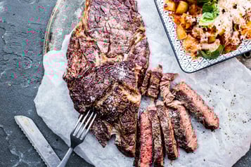 Auf einem großen mit weißem Backpapier ausgekleidetem Teller liegt ein großes Stück gebratenes Ribeye-Steak, das zum Teil in Streifen geschnitten ist. Daneben ein viereckiger gemusterter Teller mit Kartoffel-Tomaten-Würfeln garniert mit Parmesan und Basilikum.