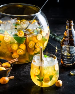 Bierbowle mit Kumquats in einem großen Glas, davor ein Glas aufgegossen, daneben ein Bitburger Winterbock. Auf dunklem Untergrund fotografiert, eine Schüssel mit Kumquats und Zimt steht im Bild.