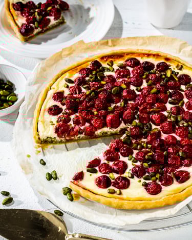 Auf einem runden Küchengitter mit hellem Backpapier befindet sich eine Tarte mit heller Creme aus der ein Stück ausgeschnitten ist, darauf sind Kirschen und Pistazien verteilt. Im linken Bildrand ist eine kleine Schüssel mit Pistazien und das geschnittene Stück der Tarte