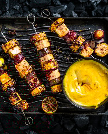 Vier marinierte Spieße mit Tofu, Mais und Möhre liegen neben leuchtend gelbem Dip.