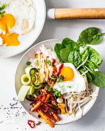 In einer Bowl sind Reis, Pilze, Spinat, Sojasprossen, Zucchini und ein Spiegelei angerichtet und mit Siracha-Sauce beträufelt.