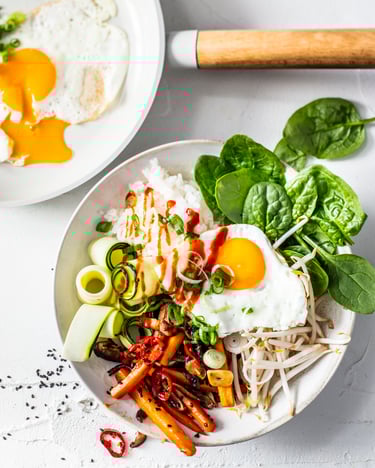 In einer Bowl sind Reis, Pilze, Spinat, Sojasprossen, Zucchini und ein Spiegelei angerichtet und mit Siracha-Sauce beträufelt.