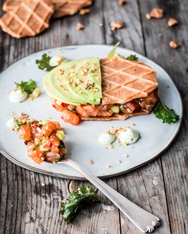 Lachstatar auf einer Gabel und zwischen Butterwaffeln mit Avocado auf einem Teller auf Holzuntergrund