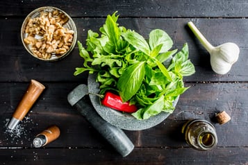 In einem grauen Mörser liegt Rucola, Basilikum und eine Chili. Daneben eine Schale mit Walnusskernen, eine Knolle Knoblauch und Öl in einer kleinen Flasche