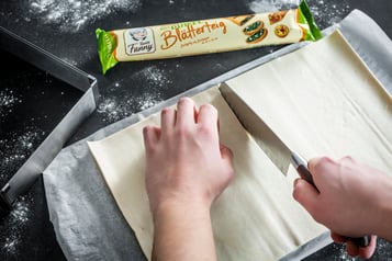 Der Blätterteig liegt auf einem schwarzen Untergrund und wird mit einem Messer halbiert