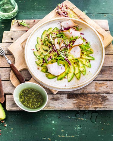 Avocado-Carpaccio liegt auf einem weißen Teller angerichtet. Darunter 2 Holzbretter und eine halbe aufgeschnittene Avocado