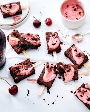 Mehrere Brownie-Stücke am Stiel liegen auf weißem Untergrund verteilt. Sie sind bestrichen mit rosanem Kirsch-Frosting und einige frische Kirschen liegen ringsherum verteilt.