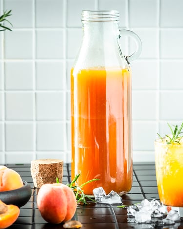 Eine große Flasche, gefüllt mit Aprikosensirup steht vor weißen Kacheln. Daneben ein Glas mit Sirup-Gemisch und einige Aprikosen liegen in einer Schale daneben.