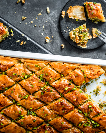 Aus einem Blech voll Baklava sind ein paar Stücke entnommen und auf einem schwarzen kleinen Teller serviert.