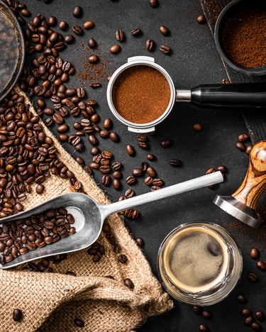 Zubehör und Zutaten für einen leckeren Espresso: Geröstete Kaffeebohnen, eine kleine Kaffeeschaufel, ein Kaffeesack, ein gefüllter Siebträger mit feinem Kaffeemehl, ein Tamper mit Holzgriff und zwei Tassen mit frisch aufgebrühtem Espresso vor dunklem Hintergrund