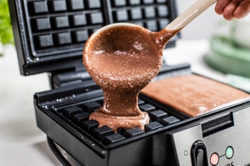 Schokoteig wird mithilfe einer Suppenkelle in ein quadratisches Waffeleisen gegeben.