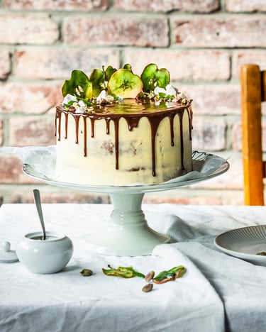 Helle Torte mit dunklem Schokoladendrip und grünen Apfelchips als Deko auf einem Tisch vor einer Backsteinwand.