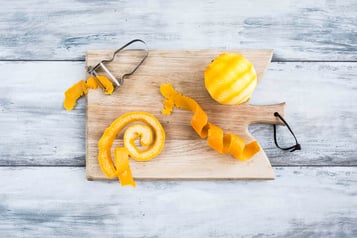 Auf einem Holzbrett liegt ein Sparschäler, eine geschälte Orange und die Orangenschale spiralförmig aufgerollt